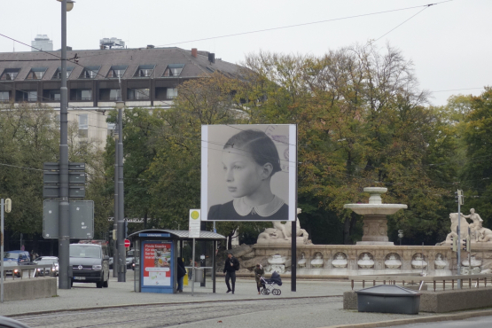 Slightly diagonal view of the billboard at Lenbachplatz. The motif shows a black and white photograph of a girl in half-profile. In the right corner appear parts of a round stamp impression, showing the imperial eagle with swastika in an oak wreath in the centre and the lettering "Polizeipräsidium München" (Munich Police Headquarters).