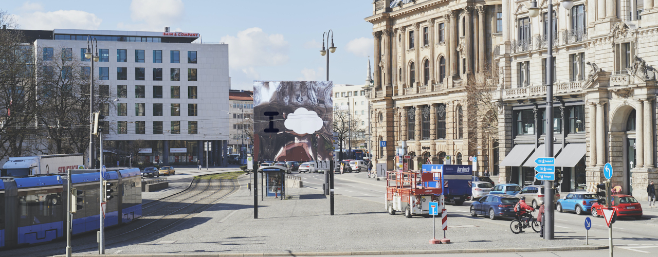 Image of the Lenbachplatz with the billboard in the middle, the row of houses on the right and a passing tram on the left. The motif is composed of a mirror with a large "I" and a cloud printed on it. The surroundings are reflected in the billboard.