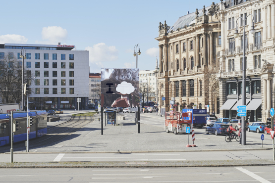 Fotografie des Lenbachplatzes mit dem Billboard in der Mitte, der Häuserreihe rechts und einer vorbeifahrenden Straßenbahn links. Das Motiv besteht aus einem Spiegel, auf dem ein großes "I" und eine Wolke aufgedruckt sind. Die Umgebung spiegelt sich im Billboard.