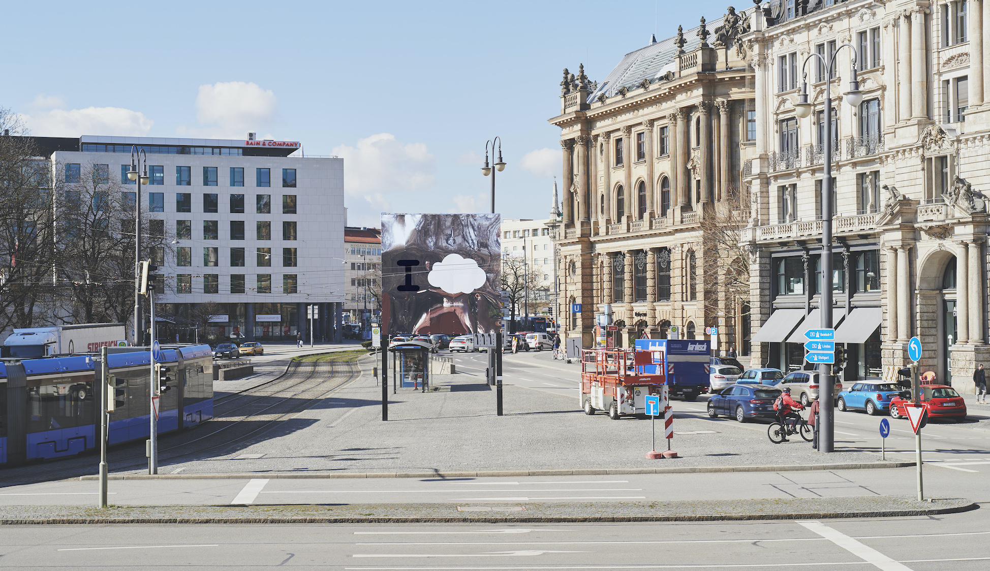 Image of the Lenbachplatz with the billboard in the middle, the row of houses on the right and a passing tram on the left.