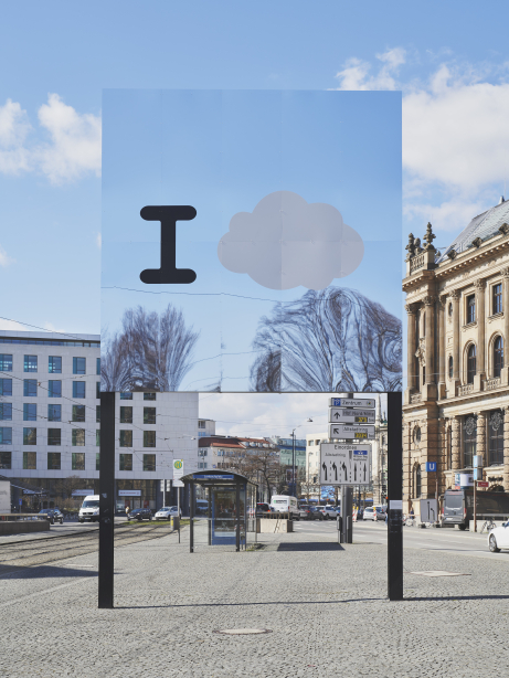 Frontalansicht des Billboards am Lenbachplatz. Das Motiv besteht aus einem Spiegel, auf dem ein großes "I" und eine Wolke aufgedruckt sind. Der Himmel und die umgebenden Bäume spiegeln sich im Billboard.