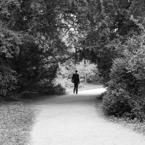 The motif shows a man in jeans and jacket, holding a pen and pad, walking on a path lined with trees. The path passes through a circular opening in the tree structure and turns to the right behind it.