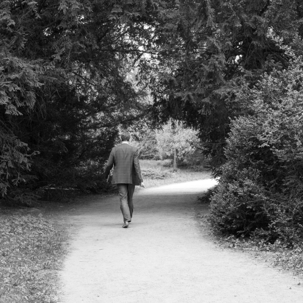 The motif shows a man in jeans and jacket, holding a pen and pad, walking on a path lined with trees. The path passes through a circular opening in the tree structure and turns to the right behind it.