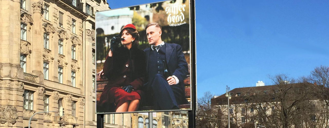 Diagonal view of the billboard. The motif shows a woman and a man sitting on a park bench, both holding a beer bottle in their hands, the woman has brought hers to her lips to drink. On the left hand appears the lettering "Sufferhead Original".