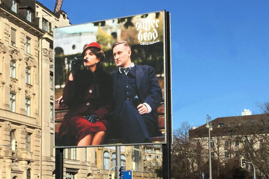 Diagonal view of the billboard. The motif shows a woman and a man sitting on a park bench, both holding a beer bottle in their hands, the woman has brought hers to her lips to drink. On the left hand appears the lettering "Sufferhead Original".