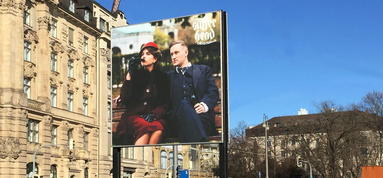 Diagonal view of the billboard. The motif shows a woman and a man sitting on a park bench, both holding a beer bottle in their hands, the woman has brought hers to her lips to drink. On the left hand appears the lettering "Sufferhead Original".