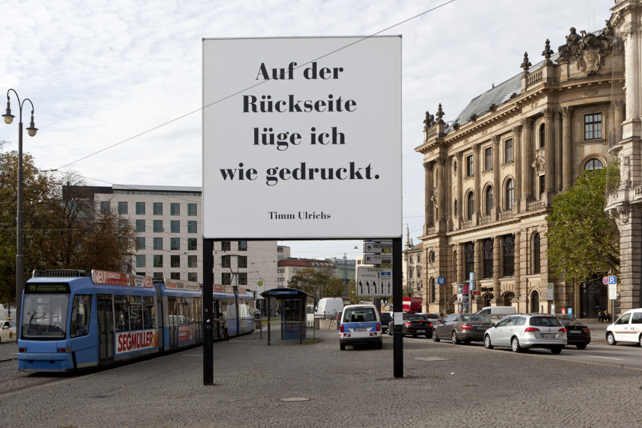 Frontal view of the billboard. To be read in large black letters on a white background: "Auf der Rückseite lüge ich wie gedruckt. Timm Ulrichs" ("I lie as printed on the reverse. Timm Ulrichs"