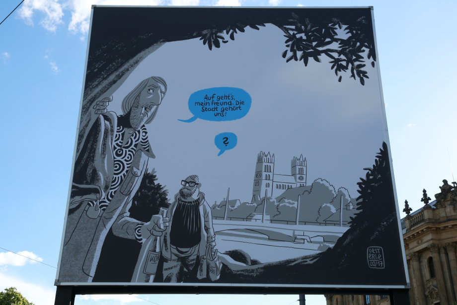 Frontal close-up view of the billboard at Lenbachplatz. The motif is executed in comic style in black and white and shows a city view of the Isar river in the background. In the foreground are two men holding cigarettes and beer bottles. A blue speech bubble next to one of the men reads: "Auf geht's, mein Freund. Die Stadt gehört uns!" ("Let's go, my friend. The city is ours!")