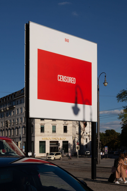Diagonale Ansicht des Billboards am Lenbachplatz. Das Billboard-Motiv zeigt ein rotes Rechteck auf weißem Grund, mittig zentriert erscheint darin das Wort "Censored" in weißer Schrift. Über dem roten Rechteck in roter Schrift zu sehen das Wort "Bad".