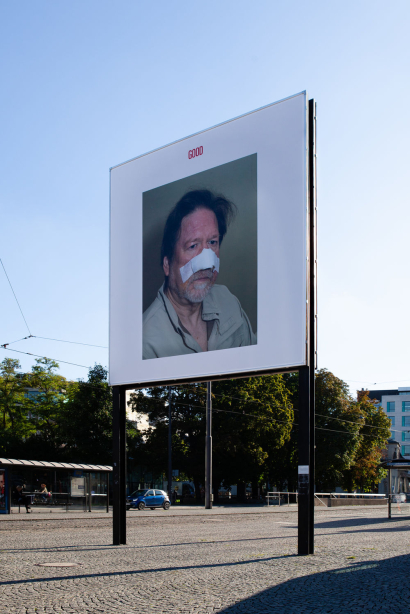 Diagonal view of the billboard at Lenbachplatz. The billboard motif shows a photo on a white background. The photo depicts a man in half-profile with a bandage over his nose and cheeks. Above this, in red lettering, is the word "Good".