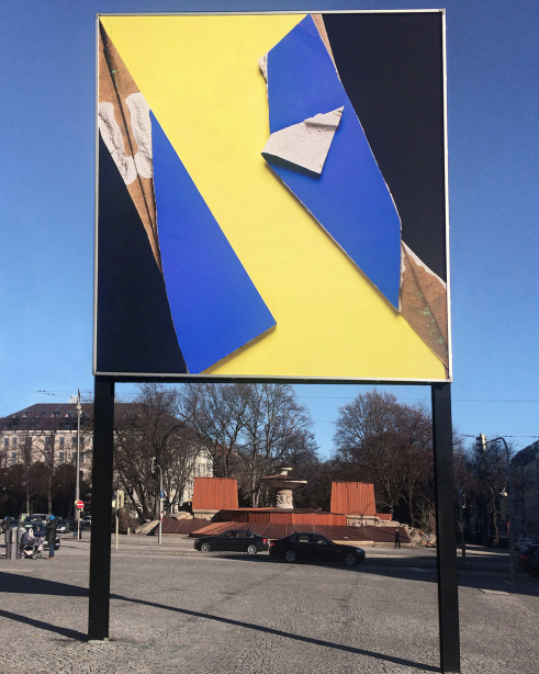 Frontal view of the billboard at Lenbachplatz. The motif shows a photograph of a plasterboard painted in yellow, blue and black. In some places, the plaster material from the inside of the board as well as the brown backing material are revealed.