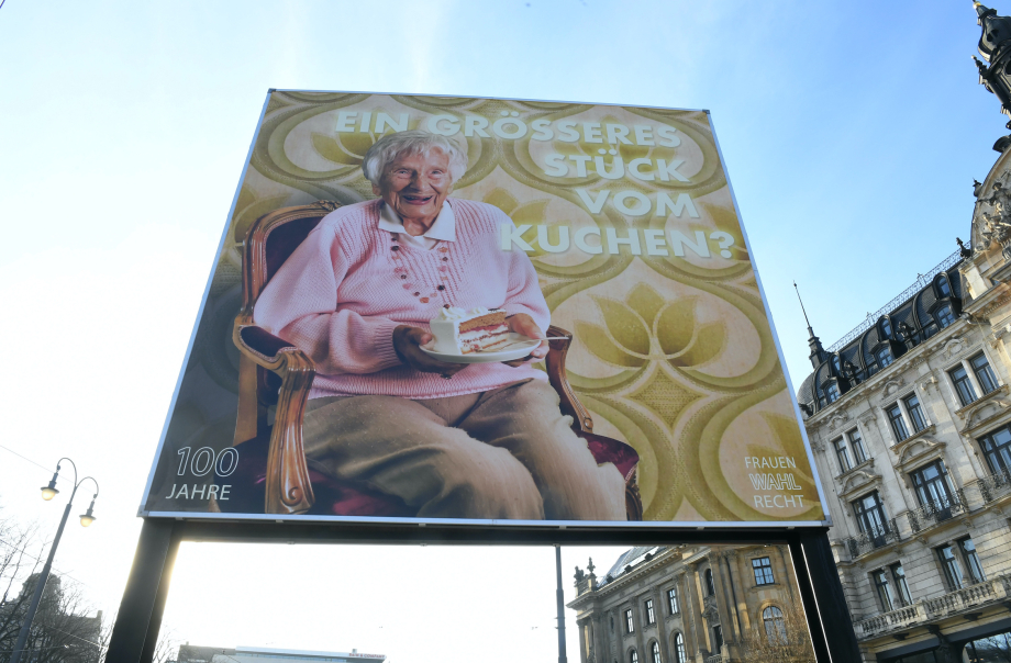 Frontal view of the billboard at Lenbachplatz. The motif shows an old lady sitting on an armchair. In her hands she holds a plate with a piece of cream cake. Above it appears the text "EIN GRÖSSERES STÜCK VOM KUCHEN?" ("A LARGER PIECE OF CAKE?").