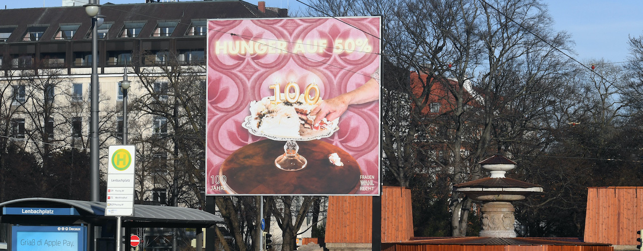 Frontal view of the billboard at Lenbachplatz. The motif shows a cream cake in the centre on a cake plate with burning birthday candles in the shape of a "100". A piece of the cake has been cut out, a hand is reaching for it. The text "Hunger auf 50%" ("Hungry for 50%") appears above it.