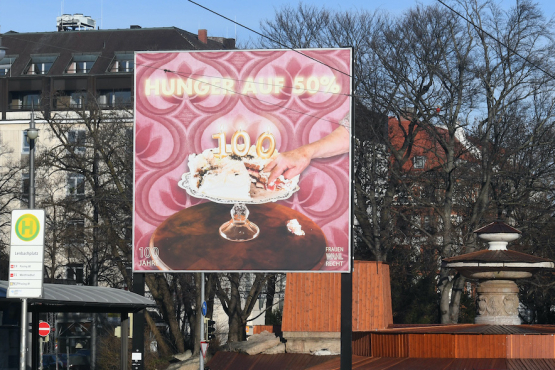 Frontal view of the billboard at Lenbachplatz. The motif shows a cream cake in the centre on a cake plate with burning birthday candles in the shape of a "100". A piece of the cake has been cut out, a hand is reaching for it. The text "Hunger auf 50%" ("Hungry for 50%") appears above it.