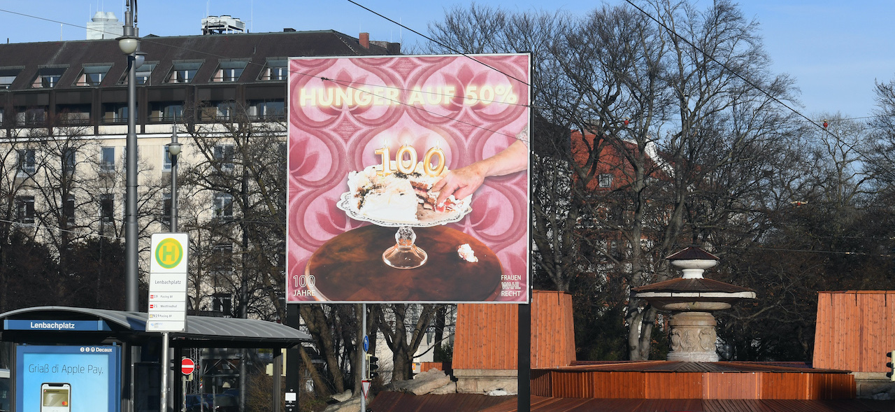 Frontal view of the billboard at Lenbachplatz. The motif shows a cream cake in the centre on a cake plate with burning birthday candles in the shape of a "100". A piece of the cake has been cut out, a hand is reaching for it. The text "Hunger auf 50%" ("Hungry for 50%") appears above it.