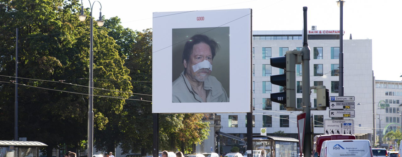 Slightly diagonal view of the billboard at Lenbachplatz. The billboard motif shows a photo on a white background. The photo depicts a man in half-profile with a bandage over his nose and cheeks. Above this, in red lettering, is the word "Good".