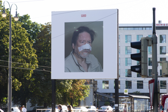 Slightly diagonal view of the billboard at Lenbachplatz. The billboard motif shows a photo on a white background. The photo depicts a man in half-profile with a bandage over his nose and cheeks. Above this, in red lettering, is the word "Good".