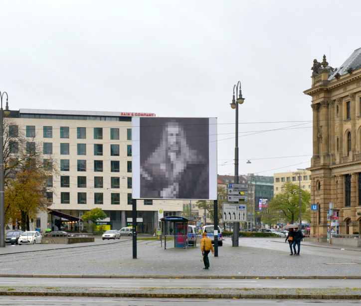 Frontal view of the billboard at Lenbachplatz. The motif shows a black and white reproduction of Albrecht Dürer's "Self-Portrait in a Fur Coat" pixelated in large squares.