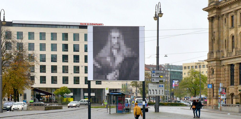 Frontale Ansicht des Billboards am Lenbachplatz. Das Motiv zeigt eine in großen Quadraten verpixelte Schwarz-Weiß-Reproduktion von Albrecht Dürers "Selbstbildnis im Pelzrock".