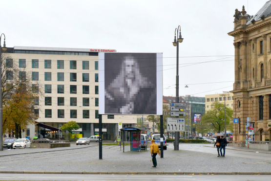 Frontal view of the billboard at Lenbachplatz. The motif shows a black and white reproduction of Albrecht Dürer's "Self-Portrait in a Fur Coat" pixelated in large squares.