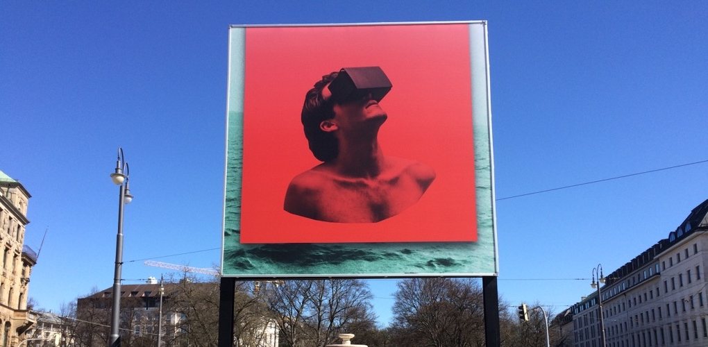 Frontal view of the billboard at Lenbachplatz. The motif shows a red square over an image of a turbulent sea. On the red background is the torso of a man wearing VR glasses looking up at the sky.