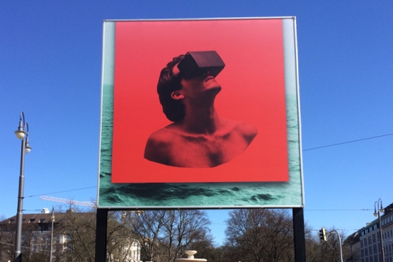 Frontal view of the billboard at Lenbachplatz. The motif shows a red square over an image of a turbulent sea. On the red background is the torso of a man wearing VR glasses looking up at the sky.