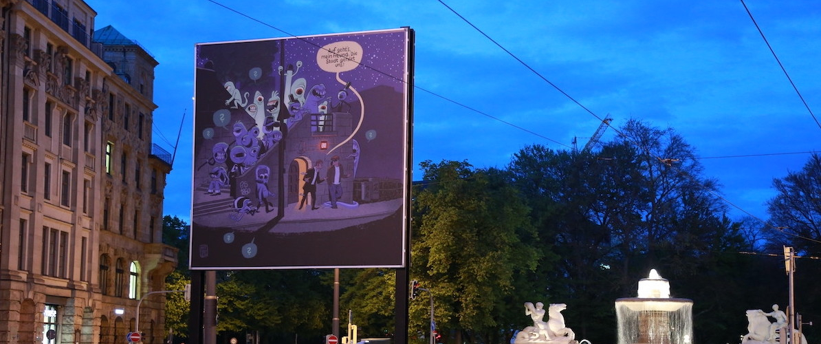 View of the billboard at Lenbachplatz in the evening hours. The motif is executed in comic style and shows two men leaving a public toilet at night. They are surrounded by a series of one-eyed monsters. A yellow speech bubble assigned to one of the man reads, "Auf geht's, mein Freund. Die Stadt gehört uns!" ("Let's go my friend. The city is ours!")