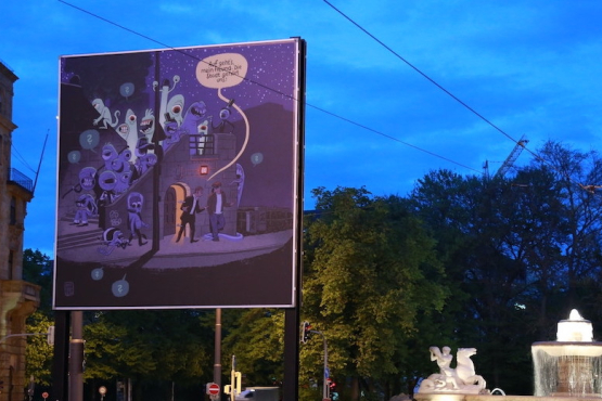 View of the billboard at Lenbachplatz in the evening hours. The motif is executed in comic style and shows two men leaving a public toilet at night. They are surrounded by a series of one-eyed monsters. A yellow speech bubble assigned to one of the man reads, "Auf geht's, mein Freund. Die Stadt gehört uns!" ("Let's go my friend. The city is ours!")