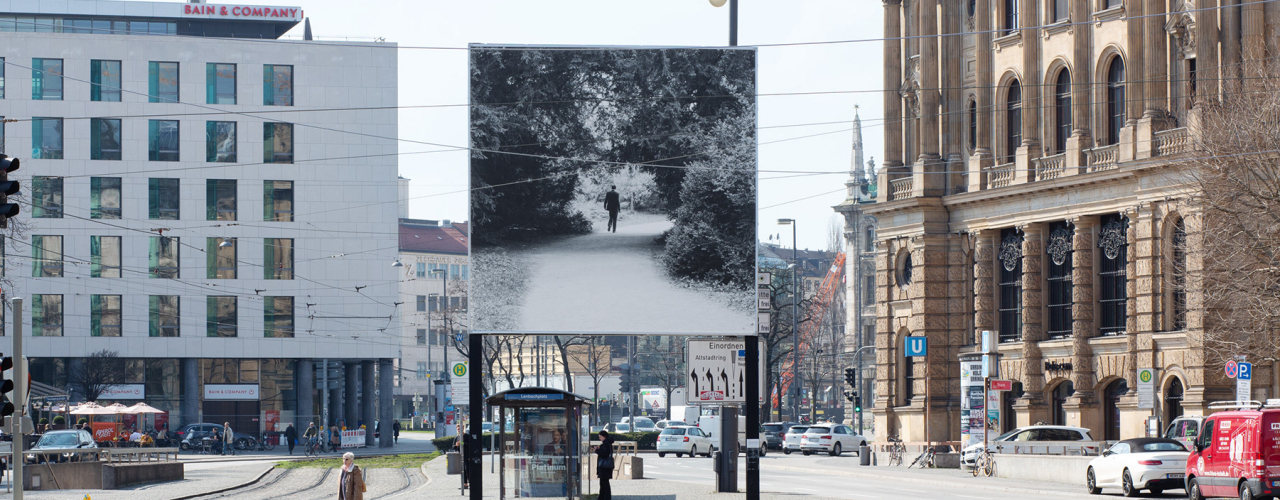 Frontal view of the side of the billboard facing away from the city. The motif shows a man in jeans and jacket, holding a pen and pad, walking on a path lined with trees. The path passes through a circular opening in the tree structure and turns to the right behind it.