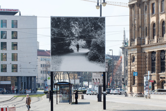 Frontal view of the side of the billboard facing away from the city. The motif shows a man in jeans and jacket, holding a pen and pad, walking on a path lined with trees. The path passes through a circular opening in the tree structure and turns to the right behind it.