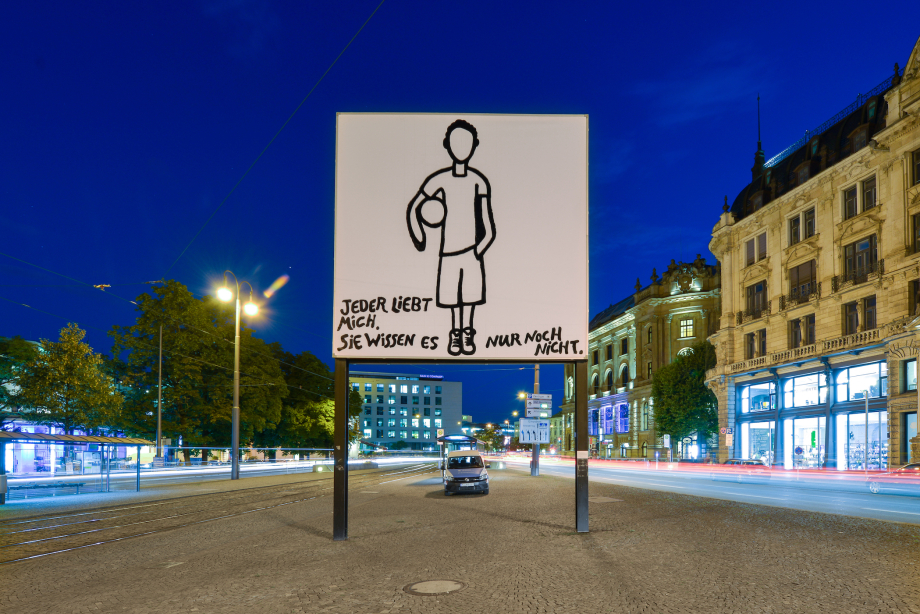 Frontal view of the billboard at Lenbachplatz in the evening hours. The motif shows a linear hand drawing of a boy standing motionless on a white background, holding a ball under his arm. The boy is depicted without a face. In the lower part of the picture appears the writing: "Jeder liebt mich. Sie wissen es nur noch nicht." ("Everyone loves me. They just don't know it yet.")
