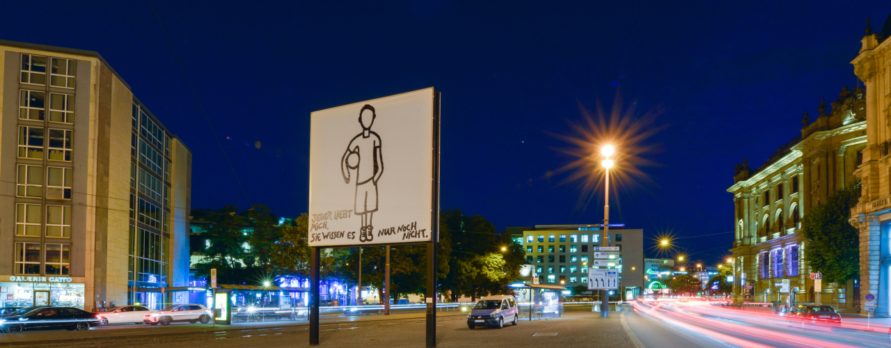 Side view of the billboard at Lenbachplatz in the evening hours. The motif shows a linear hand drawing of a boy standing motionless on a white background, holding a ball under his arm. The boy is depicted without a face. In the lower part of the picture appears the writing: "Jeder liebt mich. Sie wissesn es nur noch nicht." ("Everyone loves me. They just don't know it yet.")