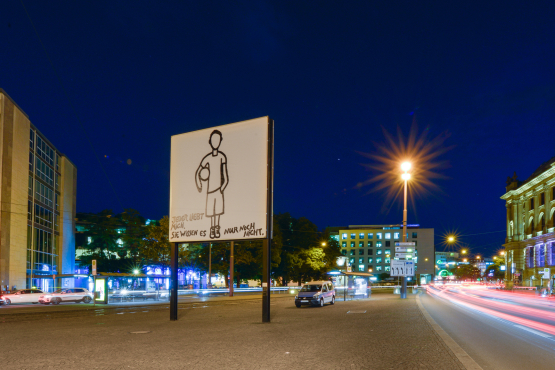 Side view of the billboard at Lenbachplatz in the evening hours. The motif shows a linear hand drawing of a boy standing motionless on a white background, holding a ball under his arm. The boy is depicted without a face. In the lower part of the picture appears the writing: "Jeder liebt mich. Sie wissesn es nur noch nicht." ("Everyone loves me. They just don't know it yet.")