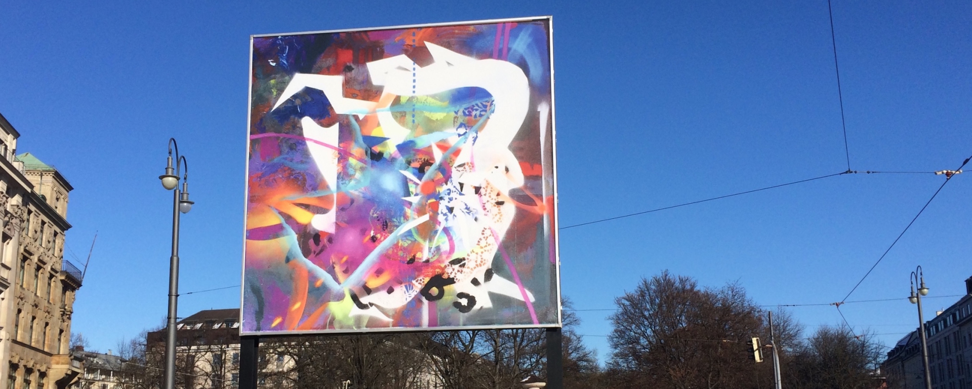 Frontale Ansicht des Billboards am Lenbachplatz. Das Motiv zeigt eine abstrakte Komposition aus leuchtend bunten, sich überlagernden Farben, Formen und Strukturen.