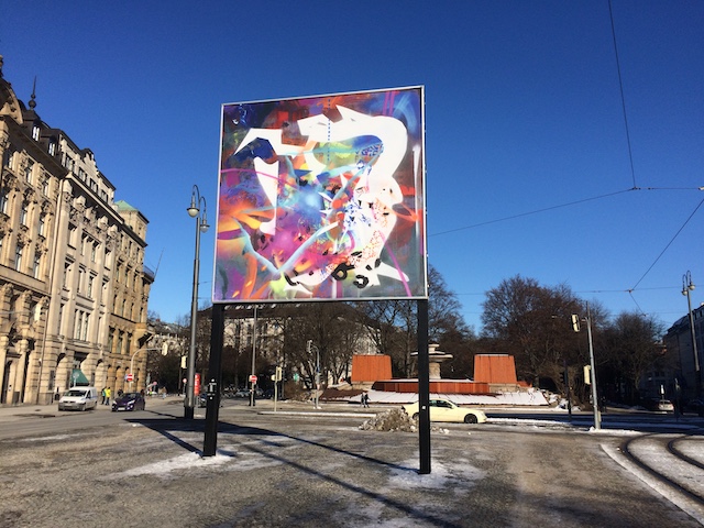 Slightly diagonal view of the billboard on Lenbachplatz. The motif shows an abstract composition of brightly colored, overlapping colors, shapes and structures.