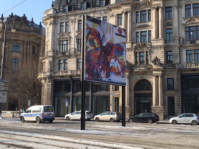 Diagonal view of the billboard on Lenbachplatz. The motif shows an abstract composition of brightly colored, overlapping colors, shapes and structures.