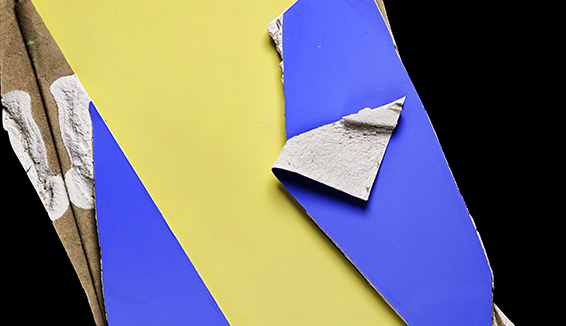 The motif shows a photograph of a plasterboard painted in yellow, blue and black. In some places, the plaster material from the inside of the board as well as the brown backing material are revealed.