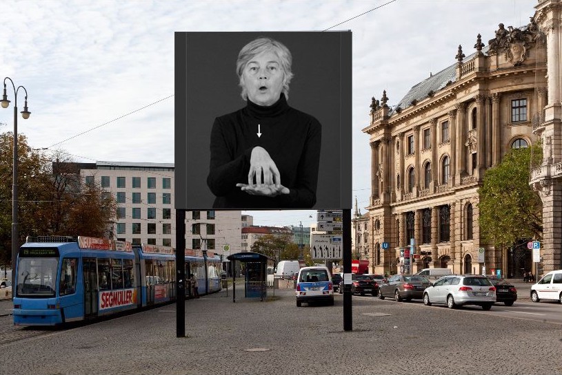 Frontal view of the billboard at Lenbachplatz. The motif shows a black and white photograph of a man in a black turtleneck jumper. His one palm points upwards, the other hand forms a closed dome above it, the sign language sign for "Karlsplatz/Stachus".