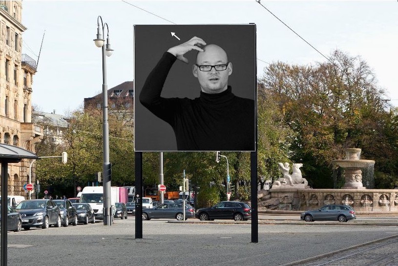 Frontal view of the billboard at Lenbachplatz. The motif shows a black and white photograph of a man in a black turtleneck jumper and glasses forming a crown on his head with one hand, the sign language sign for "king".