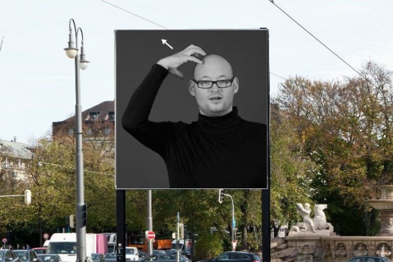 Frontal view of the billboard at Lenbachplatz. The motif shows a black and white photograph of a man in a black turtleneck jumper and glasses forming a crown on his head with one hand, the sign language sign for "king".