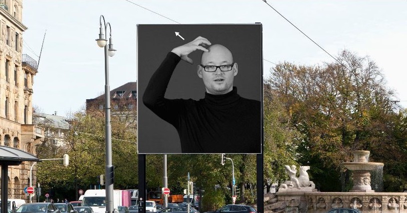 Frontale Ansicht des Billboards am Lenbachplatz. Das Motiv zeigt die Schwarz-Weiß-Fotografie eines Mannes in schwarzem Rollkragenpullover und Brille, der mit der einen Hand eine Krone an seinem Kopf formt, das Gebärdensprachezeichen für "König".