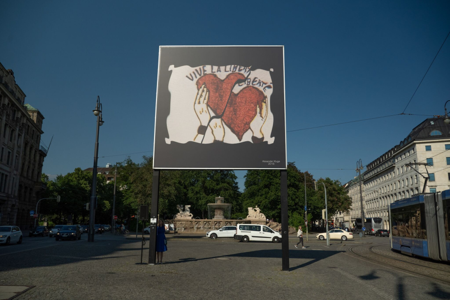 Frontal view of the billboard at Lenbachplatz. The motif shows a white rectangle with fluttering edges on a black background. Inside, hands appear holding two red hearts up, with the washed-out lettering "Vive la Liberté" above them.
