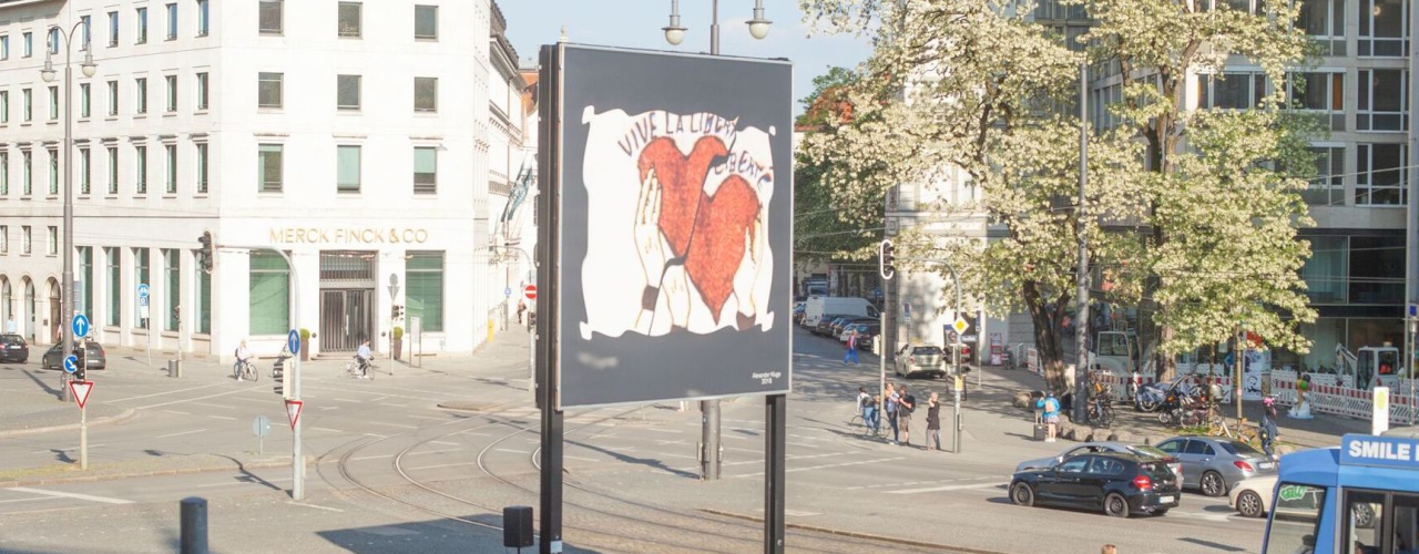 Diagnonal view of the billboard at Lenbachplatz. The motif shows a white rectangle with fluttering edges on a black background. Inside, hands appear holding two red hearts up, with the washed-out lettering "Vive la Liberté" above them.