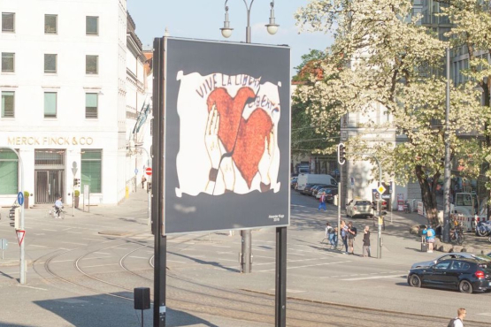 Diagnonal view of the billboard at Lenbachplatz. The motif shows a white rectangle with fluttering edges on a black background. Inside, hands appear holding two red hearts up, with the washed-out lettering "Vive la Liberté" above them.