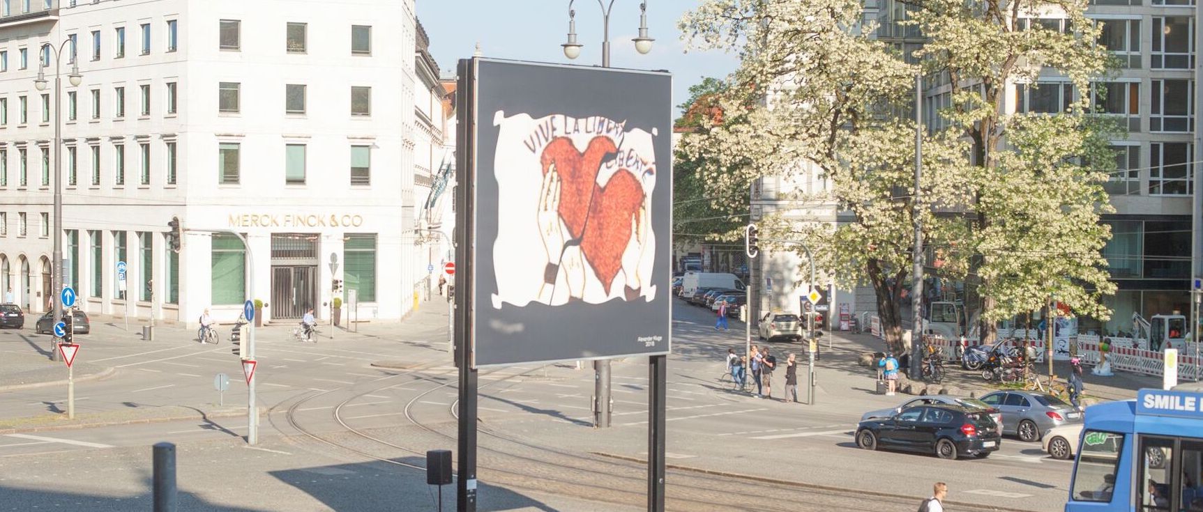 Diagnonal view of the billboard at Lenbachplatz. The motif shows a white rectangle with fluttering edges on a black background. Inside, hands appear holding two red hearts up, with the washed-out lettering "Vive la Liberté" above them.