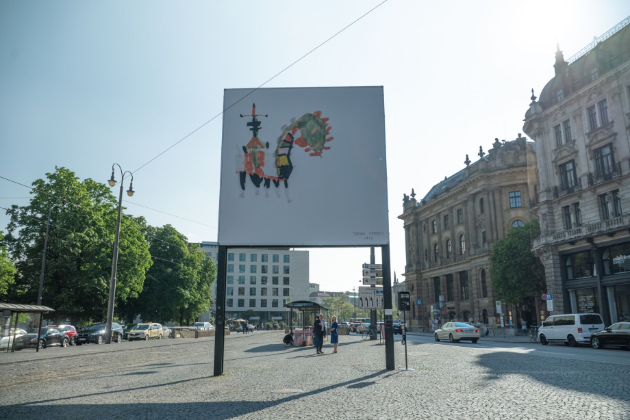 Frontale Ansicht des Billboards am Lenbachplatz. Das Motiv zeigt eine bunte Kinderzeichnung vor weißem Hintergrund, die eine Figur auf einem drachenartigen Ungeheuer reitend darstellt.