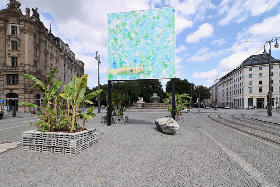 Leicht seitliche Ansicht des Billboards am Lenbachplatz umgeben von Bananenstauden. Das Motiv zeigt eine Albino-Python, die in einer abstrakten, blau-grünen Meeranmutung schwimmt. Im unteren Bildbereich erscheint in Großbuchstaben das Wort "HERE".