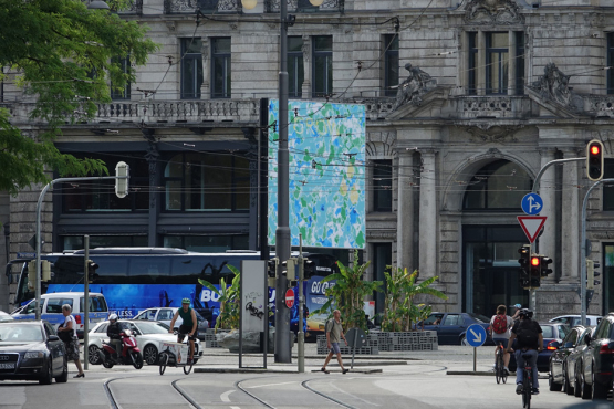 Seitliche Ansicht des Billboards am Lenbachplatz aus der Ferne in der Rushhour. Das Motiv zeigt eine abstrakte, blau-grüne Meeranmutung. Im oberen Bildbereich erscheint in Großbuchstaben das Wort "GROW".