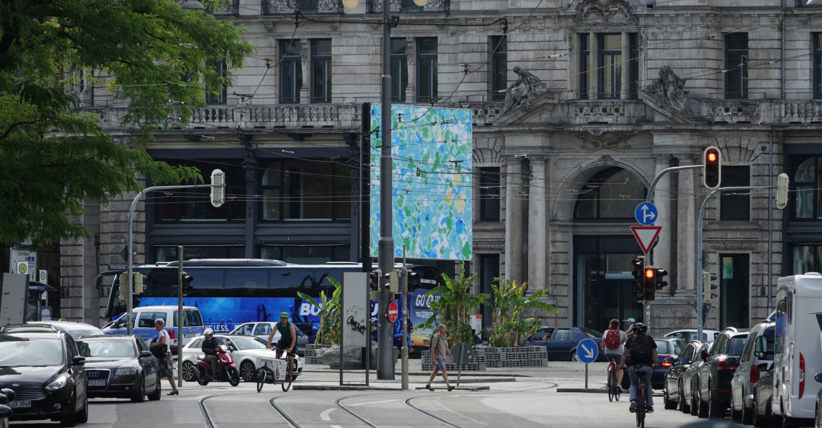 Side view of the billboard at Lenbachplatz from a distance during rush hour. The image shows an abstract blue-green sea. The word "GROW" appears in capital letters at the top of the image.