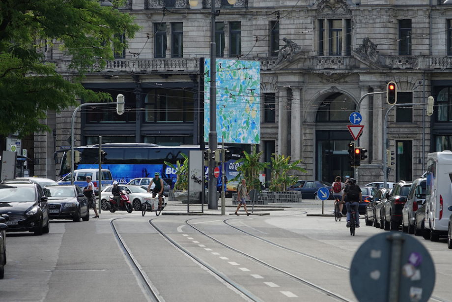 Side view of the billboard at Lenbachplatz from a distance during rush hour. The image shows an abstract blue-green sea. The word "GROW" appears in capital letters at the top of the image.